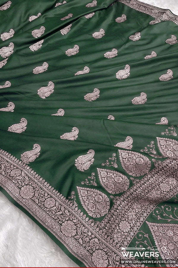 A bottle green Banarasi silk saree with intricate silver paisley and floral zari embroidery, featuring a wide decorative border and a matching dupatta draped over a white surface.