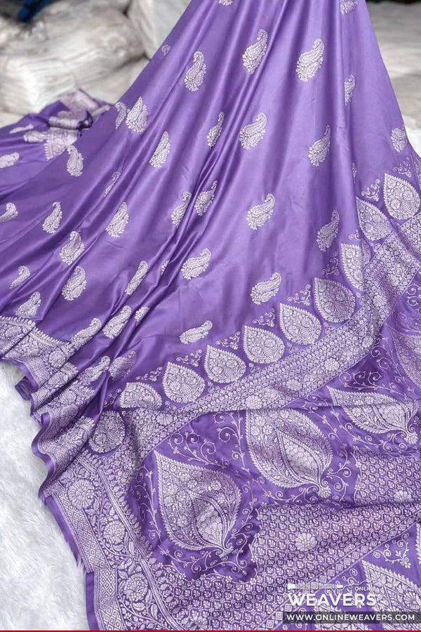 A lilac purple Banarasi silk saree with intricate silver paisley and floral motifs, draped elegantly on a white textured surface. A watermark for 'Weavers Online' is visible in the bottom right corner.