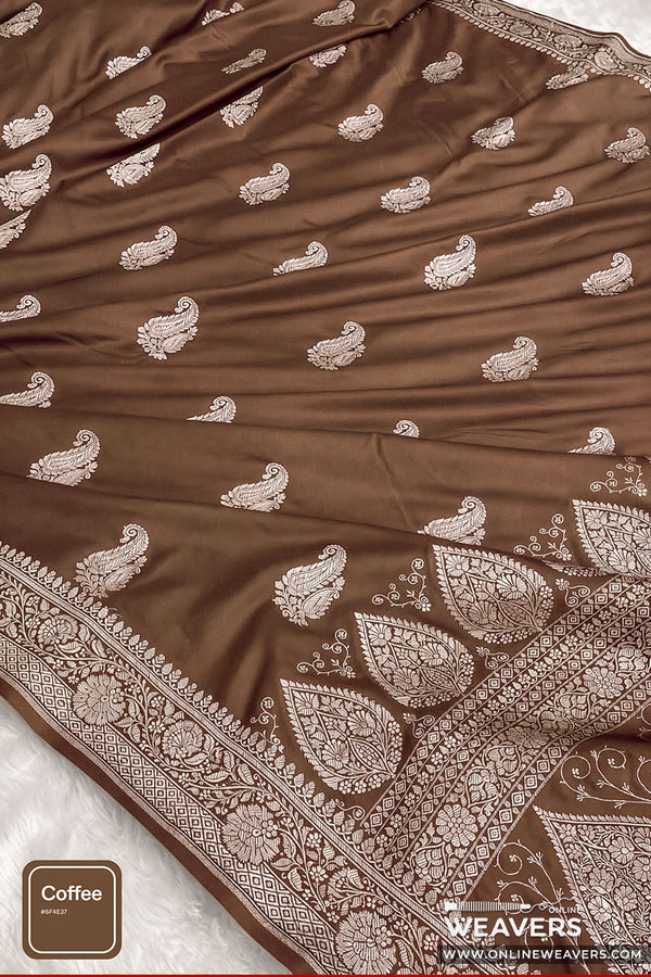 A coffee brown color Banaarsi silk saree with intricate silver paisley and floral motifs, featuring a wide decorative border, laid out on a white textured surface.