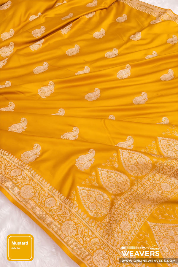 A mustard yellow Banarasi Silk Saree, featuring woven cream paisley and floral motifs with a detailed cream border. A yellow label in the bottom-left corner reads 'Mustard Yellow' and the bottom-right corner features a 'OnlineWeavers' brand logo.