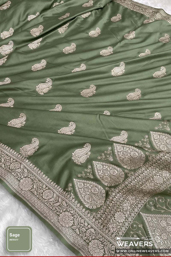 An olive sage green Banarasi silk saree with intricate silver paisley and floral motifs, featuring a wide decorative silver border, shown draped on a white textured surface.