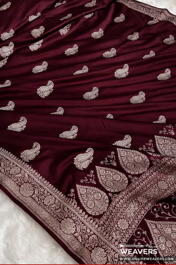 A close-up of a deep maroon silk Banarasi saree with intricate silver Banarasi paisley and border embroidery, laid flat on a white textured surface.