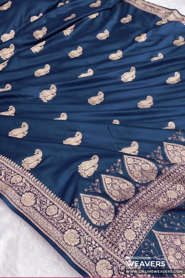 A peacock blue Banarasi silk mashru saree featuring intricate gold zari small paisley motifs and a heavily decorated border, laid flat on a white surface.