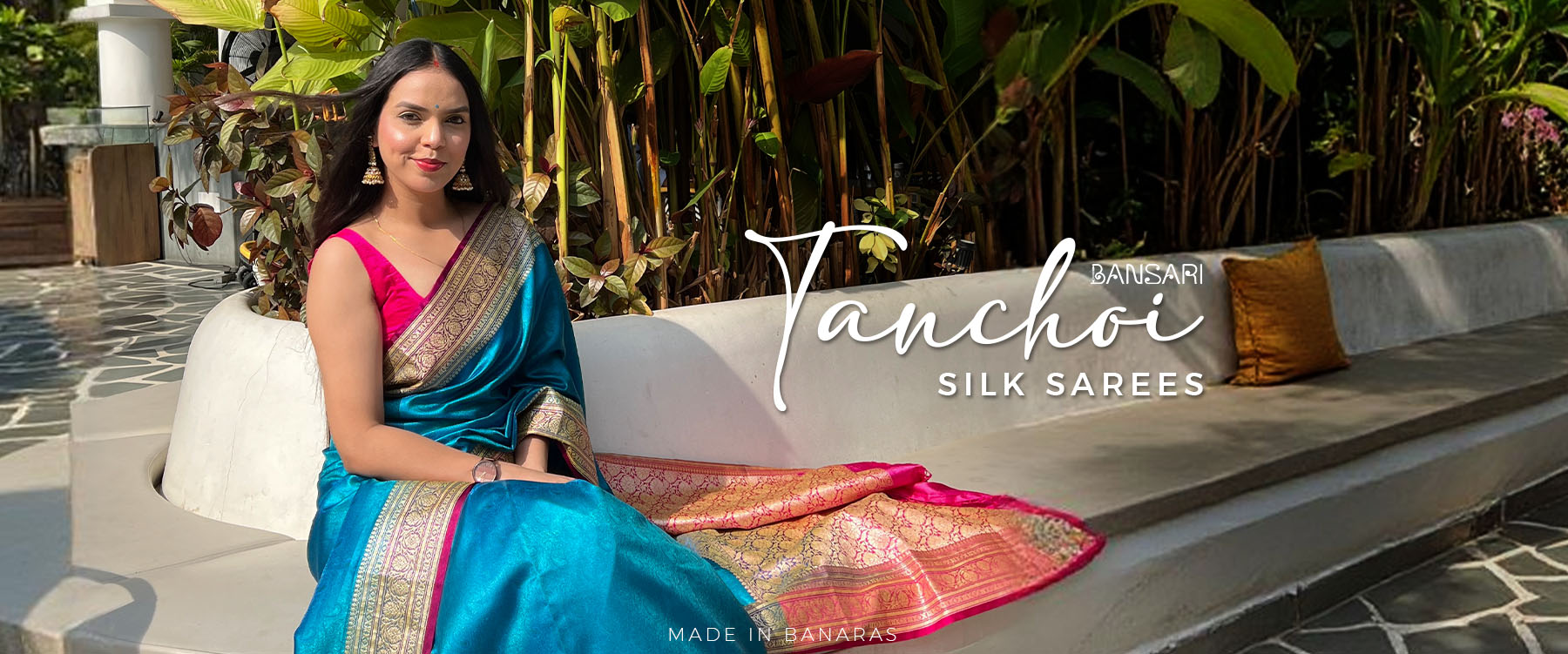 Woman in a tanchoi blue saree sitting on a bench with 'Banarasi Saree manufacturer' branding.