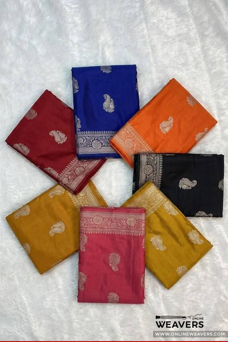 Seven folded Banarasi silk sarees in various colors (red, blue, orange, black, mustard yellow, pink, and beige) are arranged in a circular pattern on a white textured fabric background. Each saree features a gold zari paisley motif and border detailing.