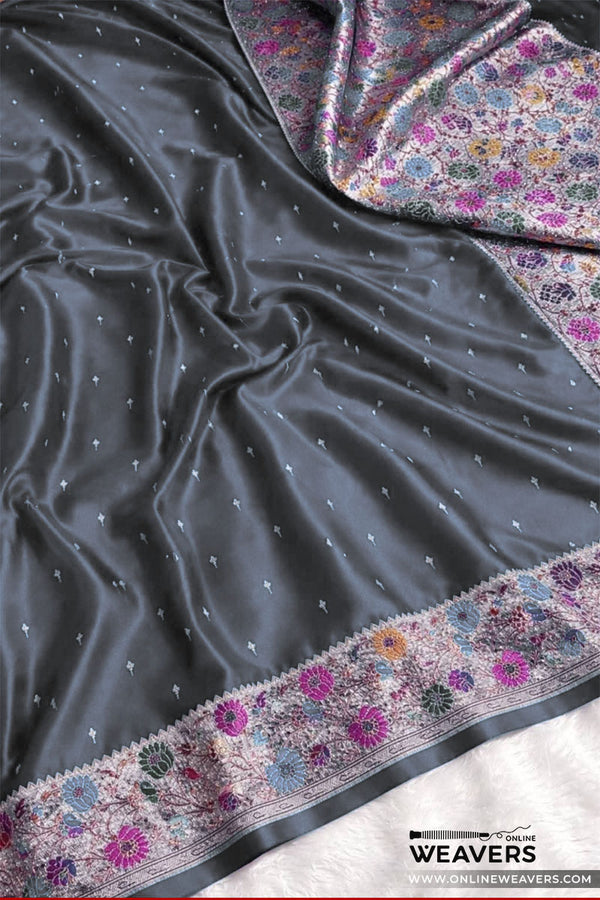 A dark gray mashru silk saree drape with small metallic dot motifs and a heavily embroidered border in pink, purple, blue, and gold floral patterns, shown on a white surface with a OnlineWeavers watermark.