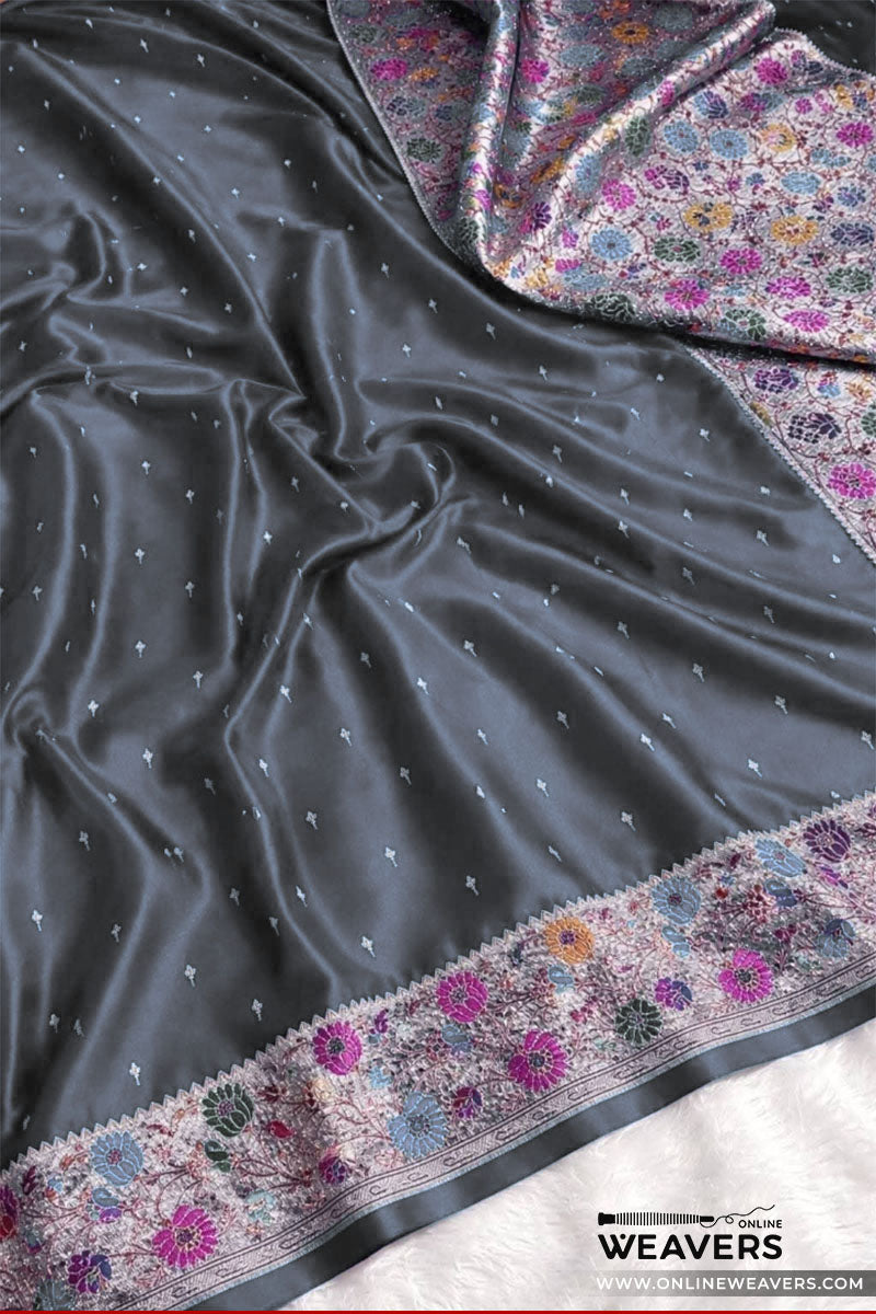 A dark gray mashru silk saree drape with small metallic dot motifs and a heavily embroidered border in pink, purple, blue, and gold floral patterns, shown on a white surface with a OnlineWeavers watermark.