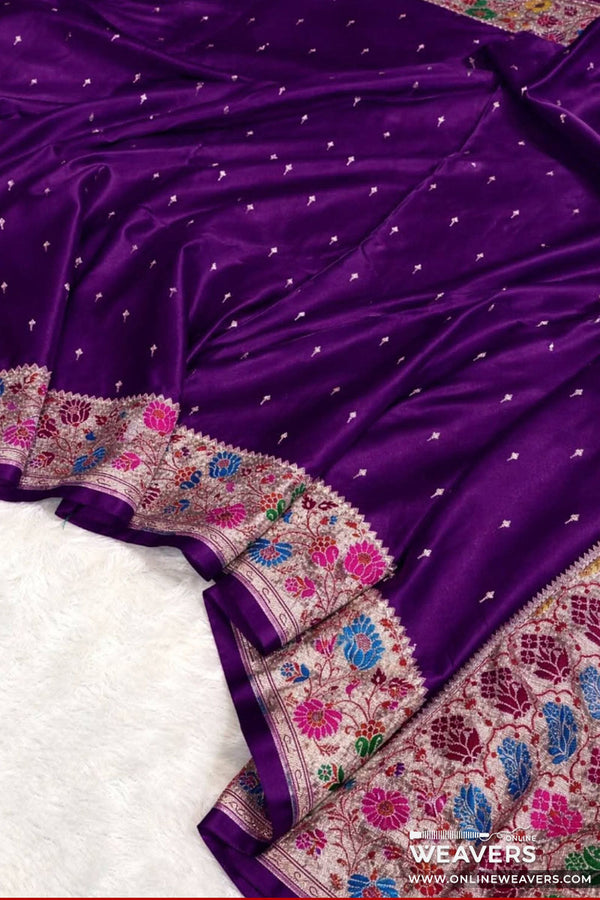 A close-up of a purple mashru silk Banarasi saree with small metallic motifs and a heavily embroidered border in pink, blue, and gold, laid over a white rug. With 'OnlineWeavers' branding